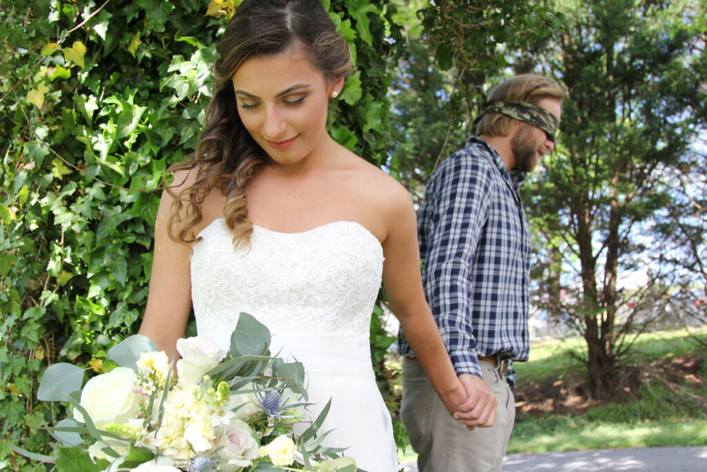 Wedding photography of bride holding a bouquet and her blindfolded husband's hand.